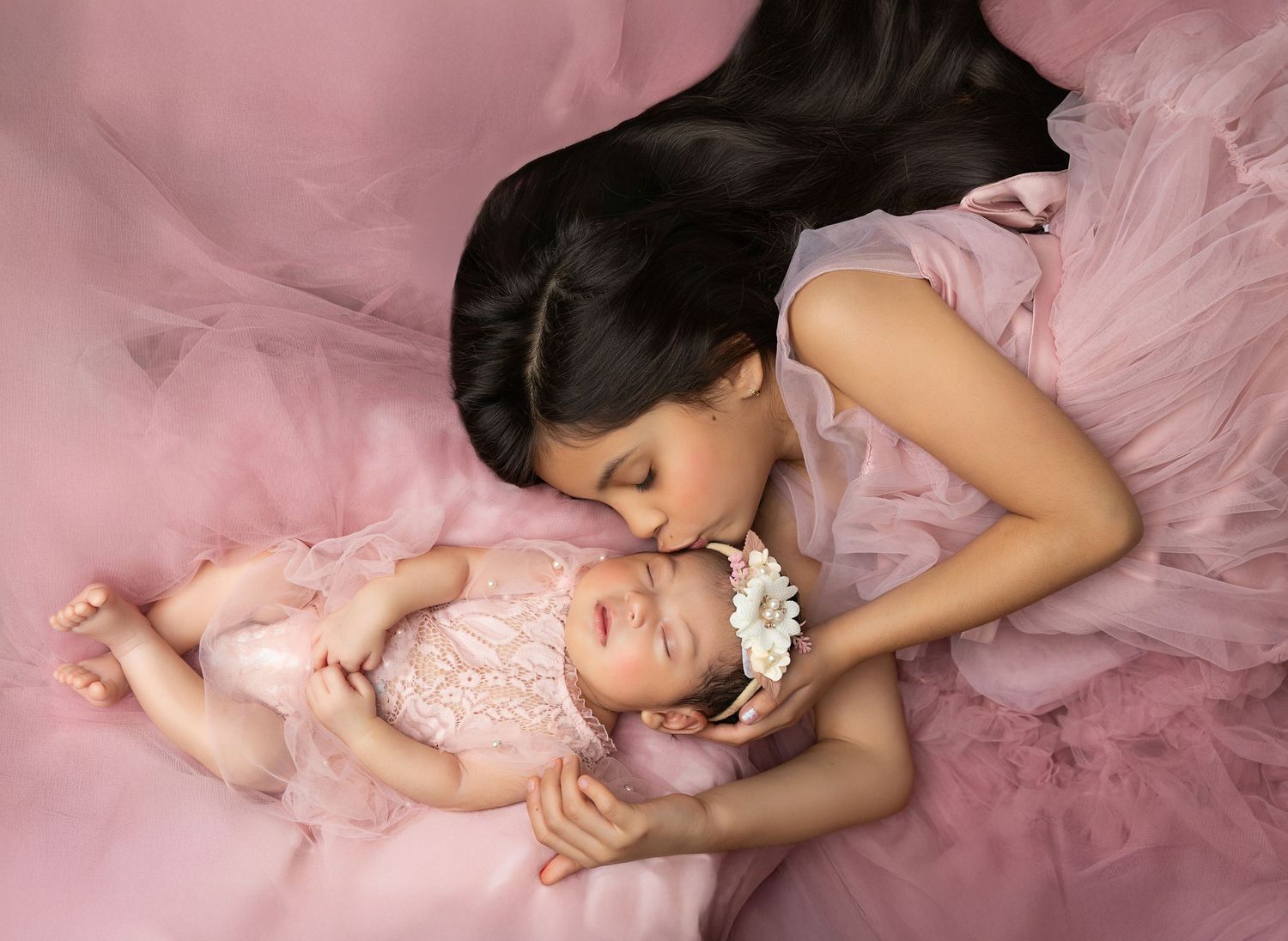 A parent and baby lie together on pink tulle fabric in matching pink outfits for a tender portrait session.
