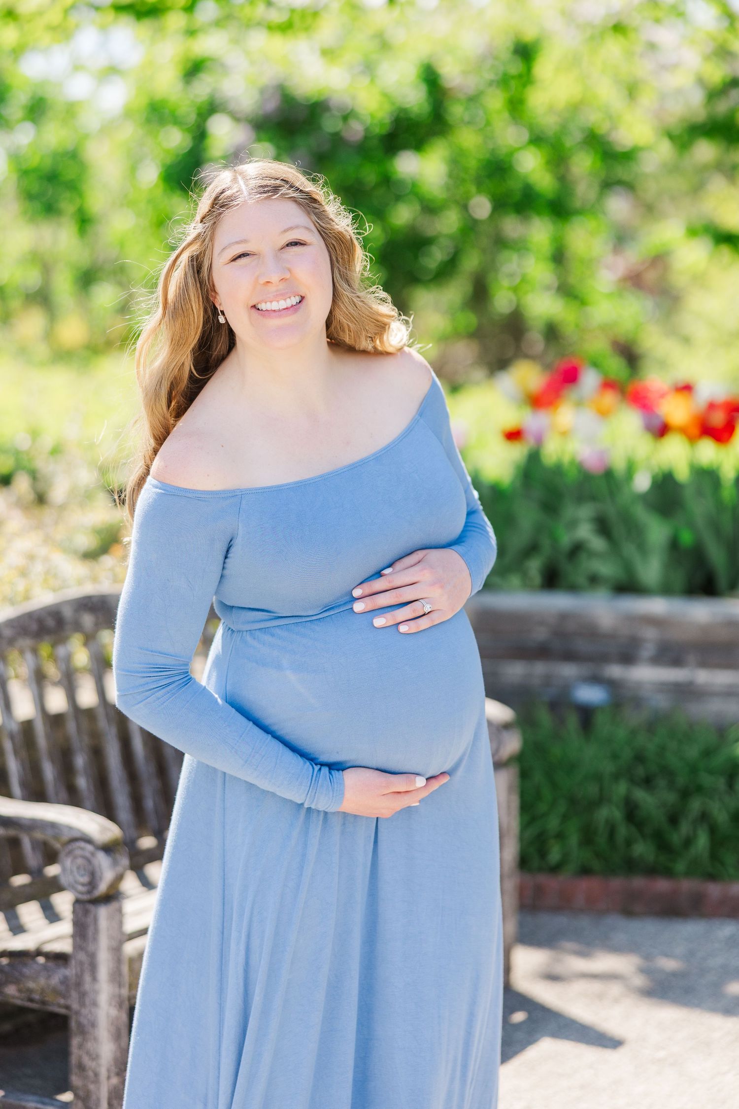 Pregnant person wearing light blue off-shoulder maxi dress poses outdoors in garden setting with red tulips and greenery at Chicago Botanic