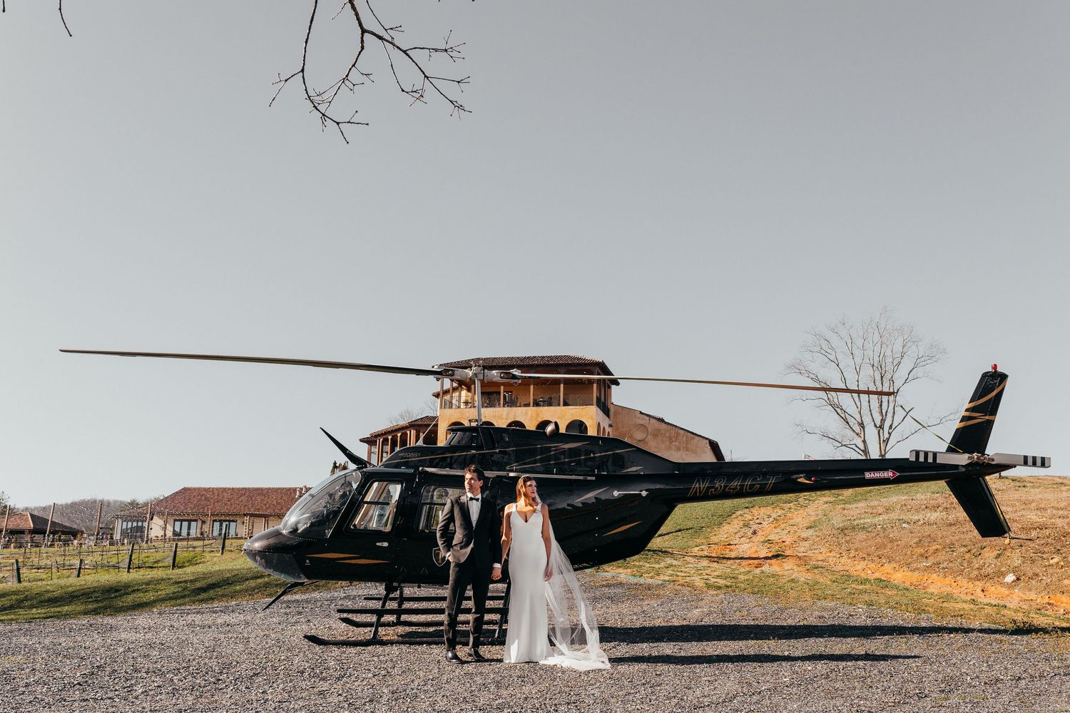 A couple stands in front of a sleek black helicopter on a gravel driveway at a rustic venue.