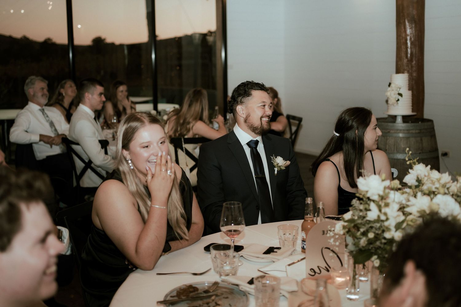Wedding reception guests enjoying speeches and dinner in a rustic venue.