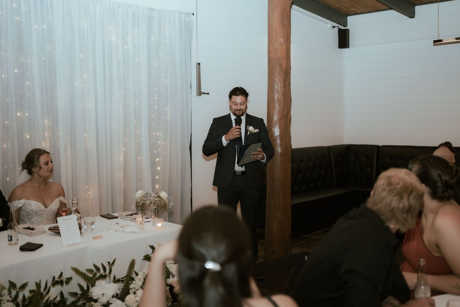 Speech being delivered during wedding reception with white backdrop and mood lighting.