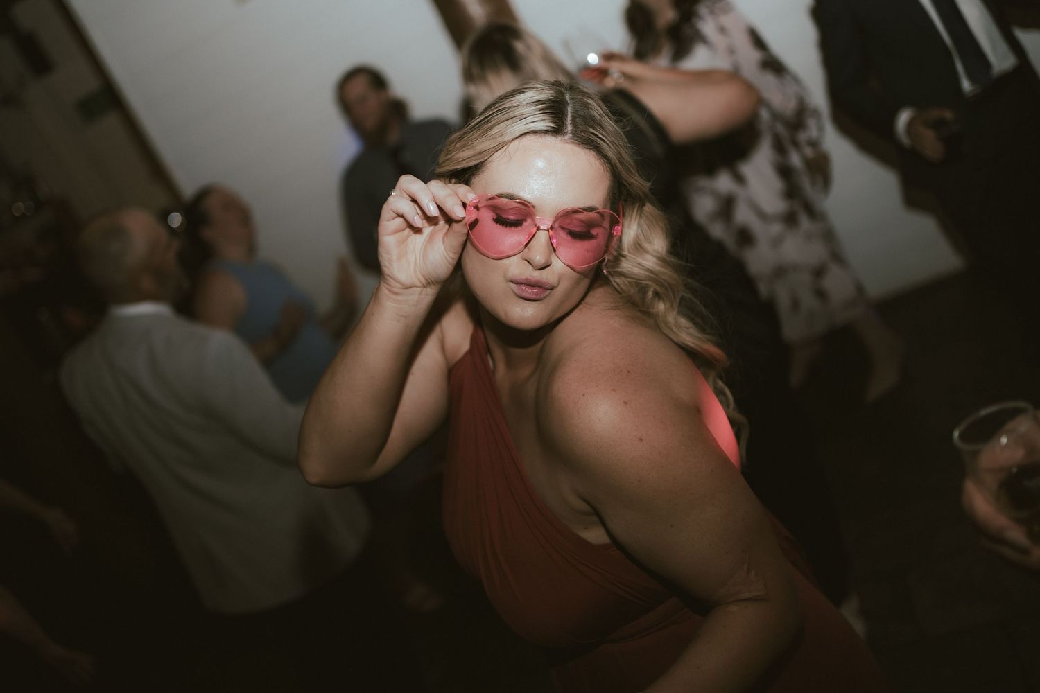 Person adjusting pink sunglasses on dance floor with crowd in background.