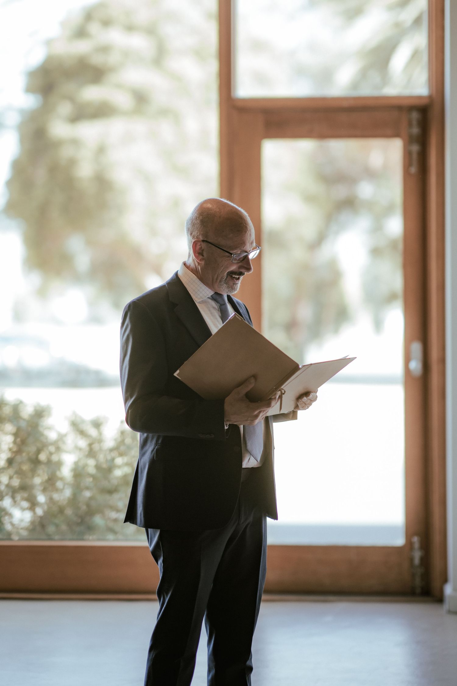 Person in formal attire reading from papers near large windows.