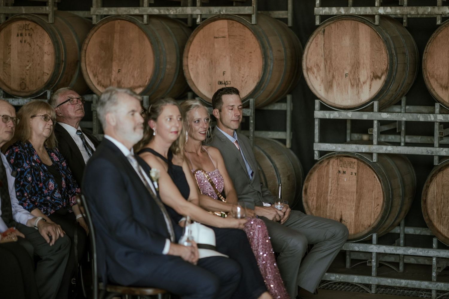 Wedding guests seated in rows at a winery ceremony with large wooden barrels in the background.