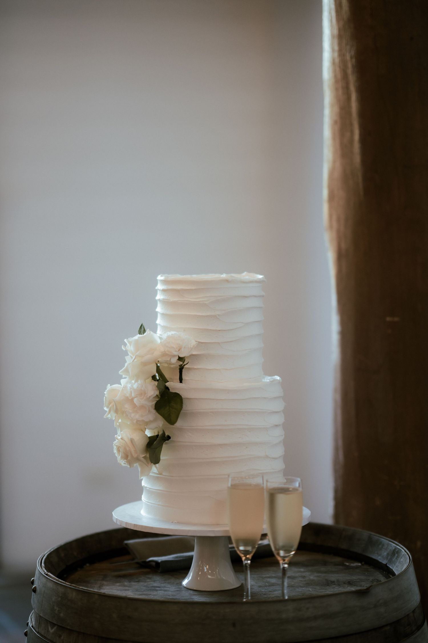 Elegant white tiered wedding cake with flowers and champagne glasses on rustic barrel.