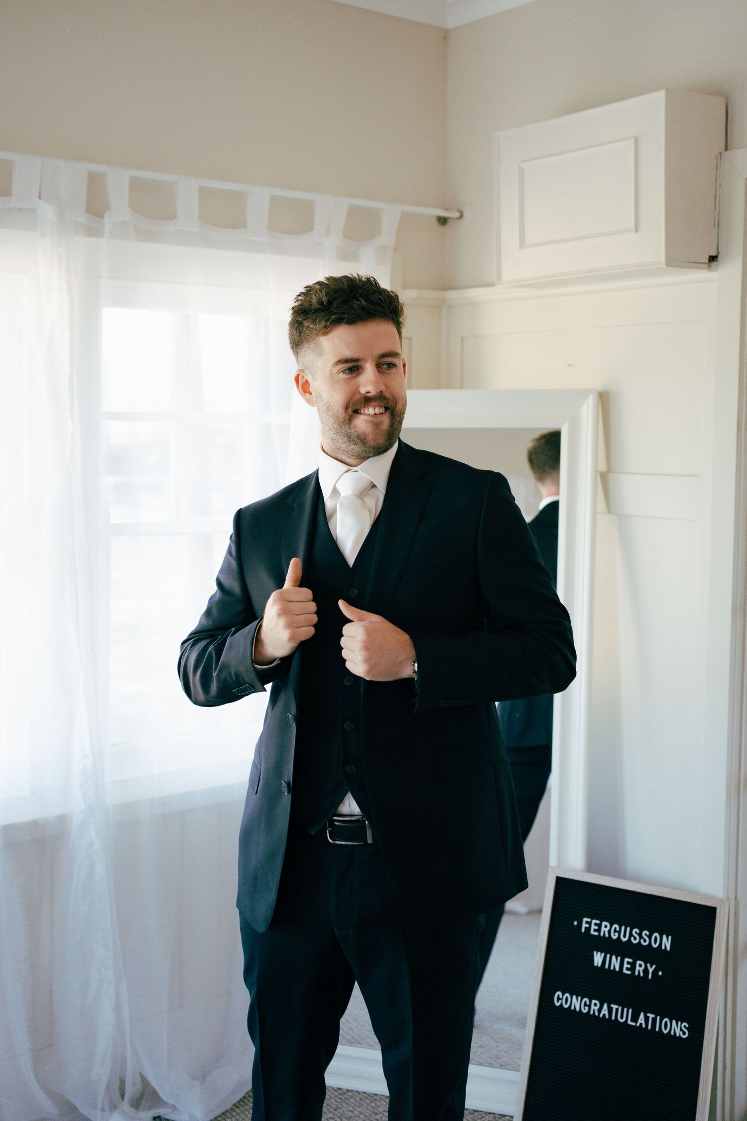 Groom in classic black tuxedo adjusts his jacket while smiling in a bright room.