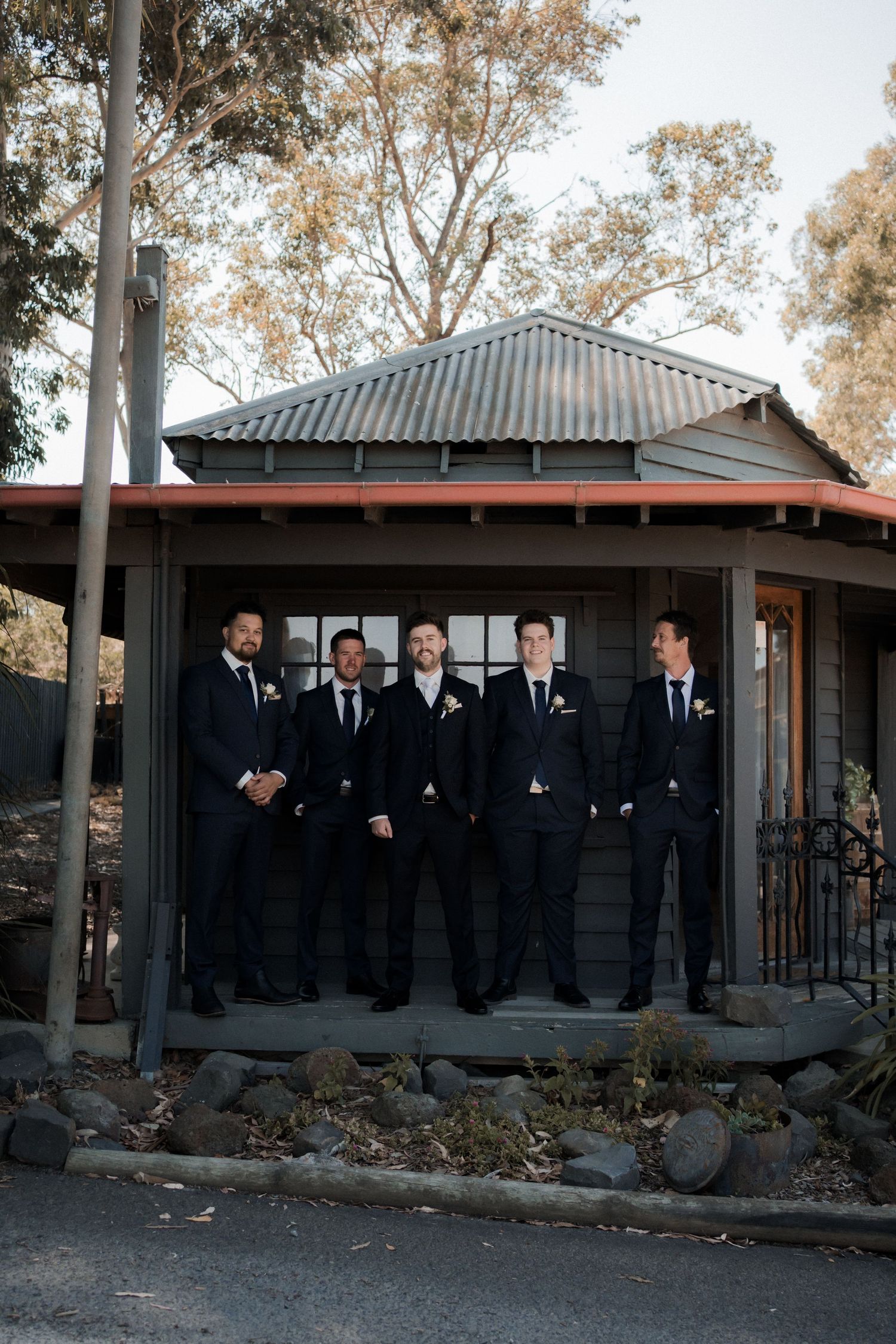 Groomsmen in formal attire lined up for photo on stone porch of rustic building.