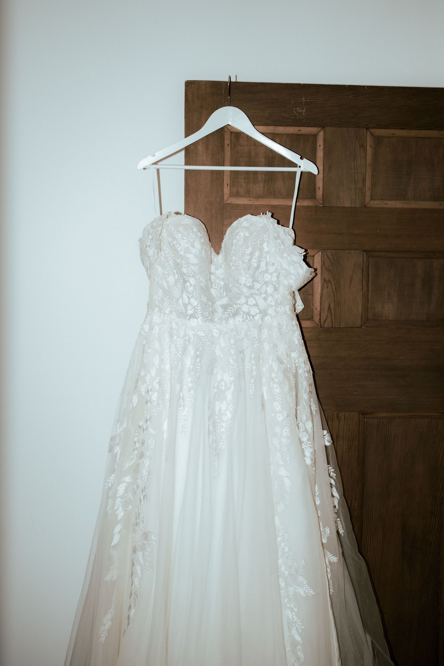 Elegant white lace wedding dress hanging on white hanger against wooden door.