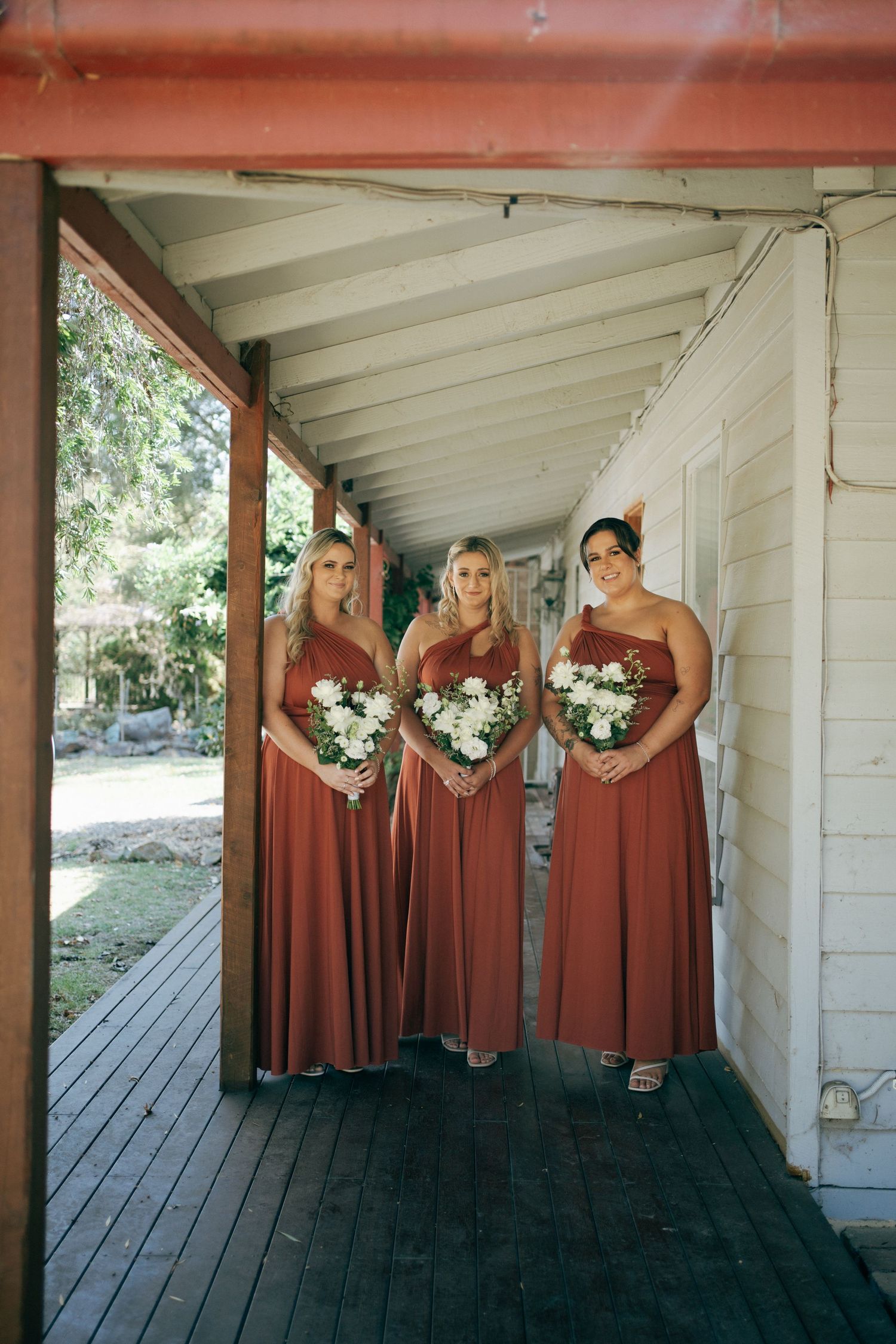 Three bridesmaids in rust-colored dresses holding white bouquets pose on wooden porch.