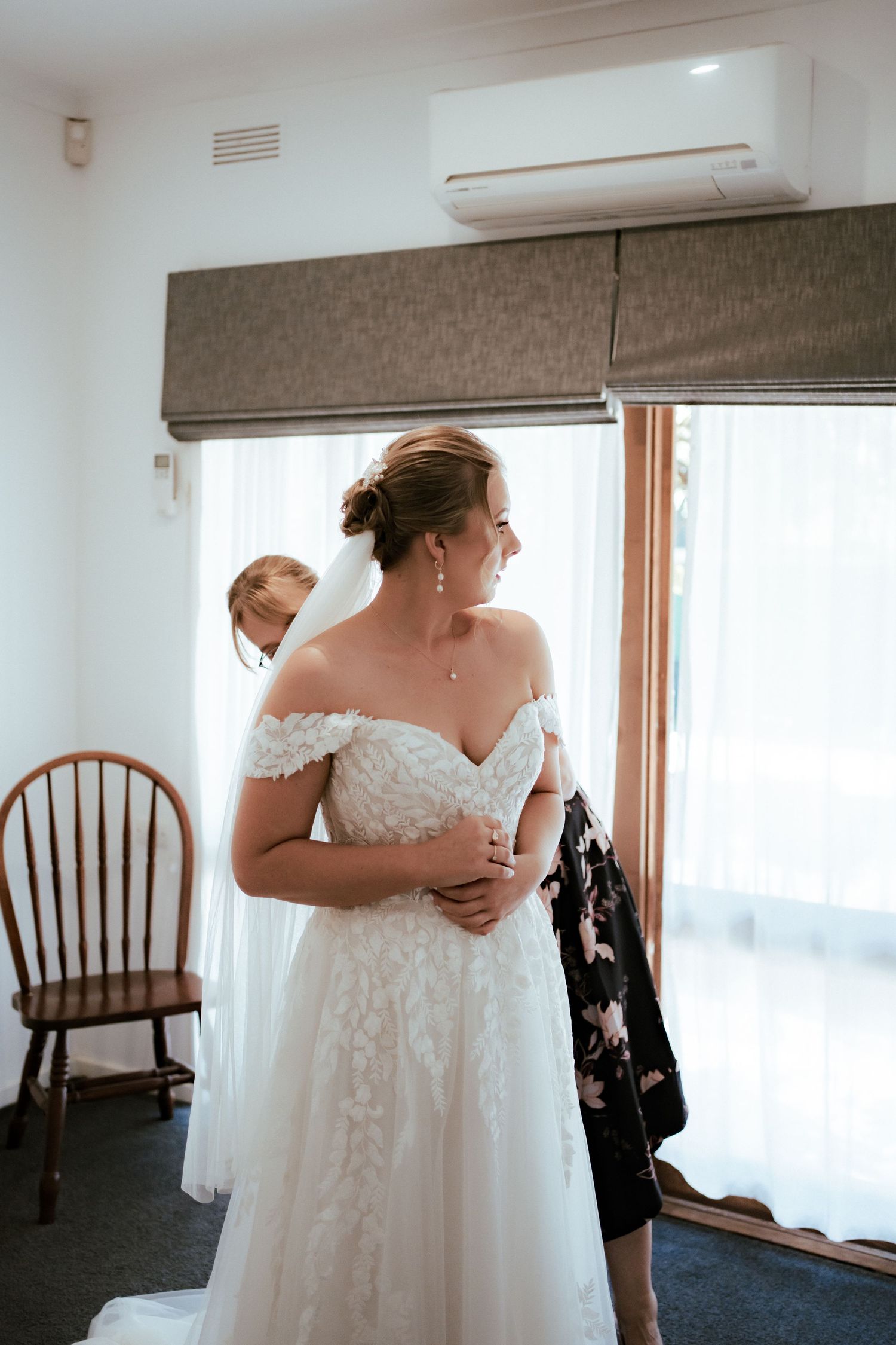 A bride in an off-shoulder lace wedding gown stands in a room with a wooden chair and window with brown roman shade.