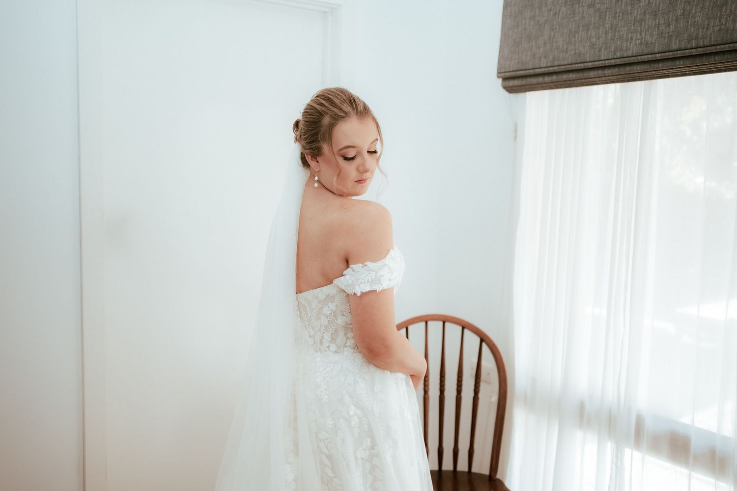Back view of a bride wearing an off-shoulder lace wedding dress near a window and wooden chair.