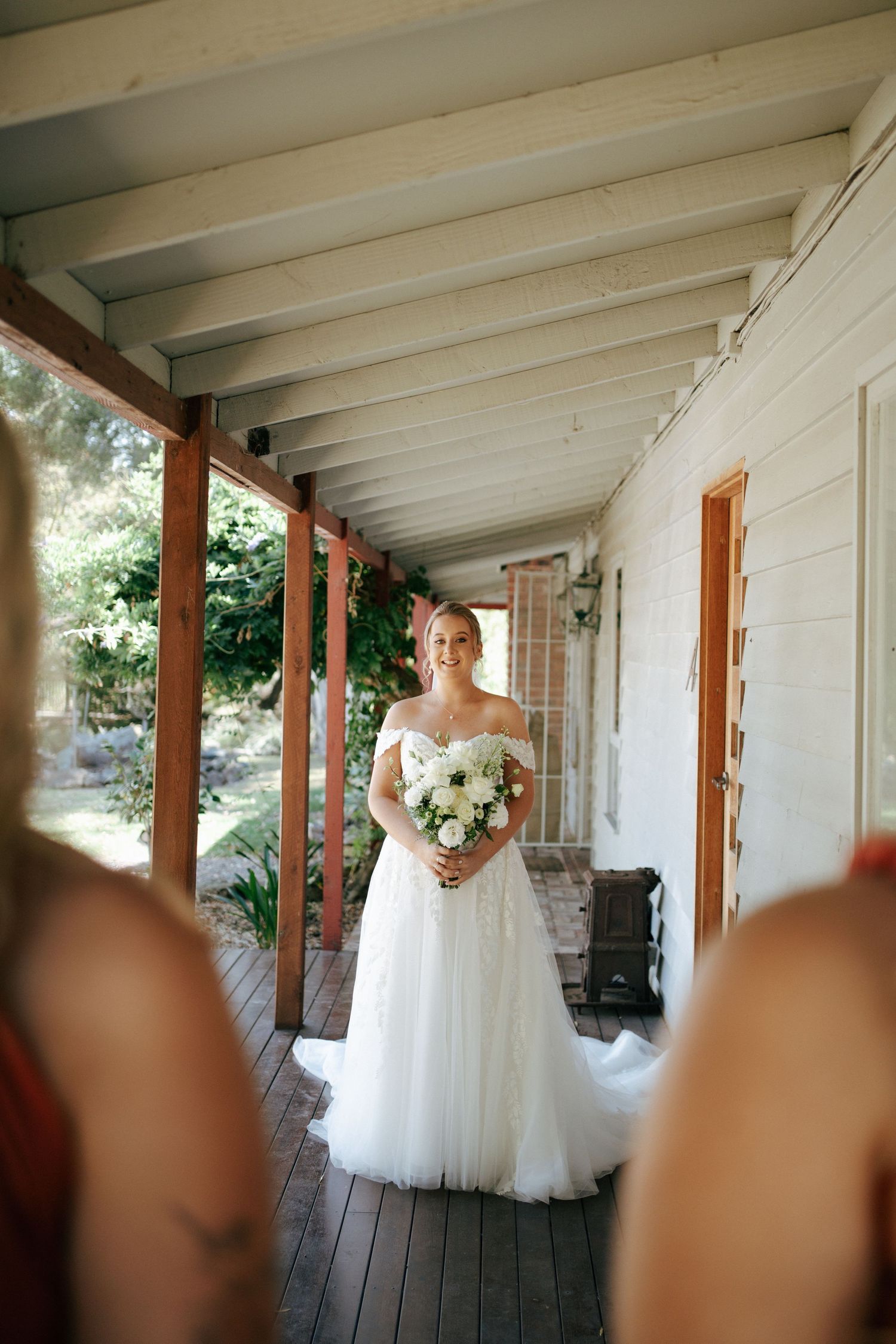 A bride in a white tulle gown holds a bouquet while standing on a rustic covered porch.