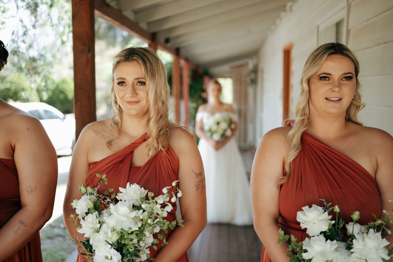 Bridesmaids in rust-colored one-shoulder dresses hold white bouquets on a covered porch.