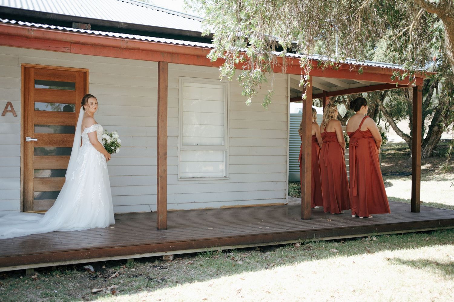 Wedding party in rust dresses and white gown pose on wooden deck of country house.