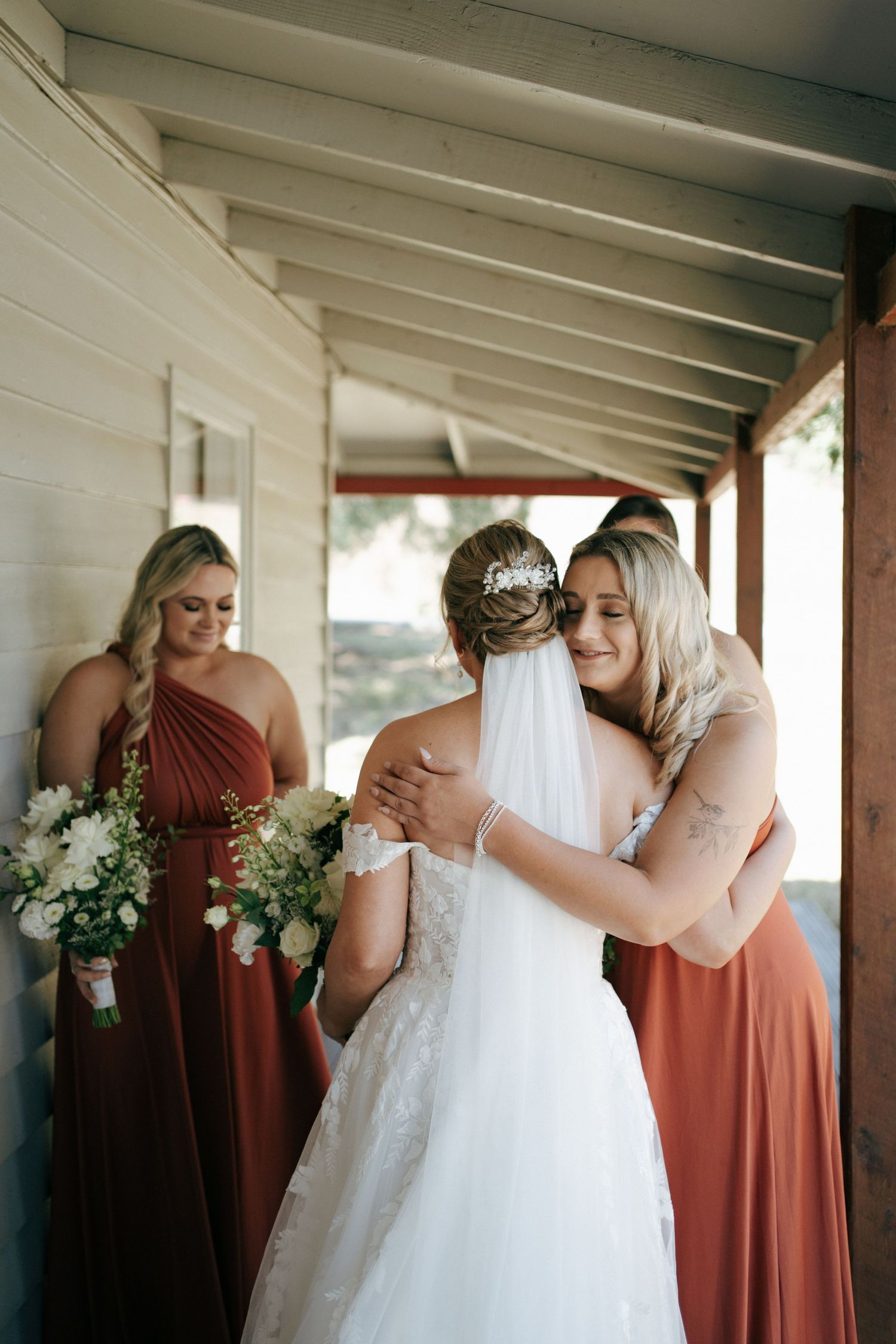 Emotional wedding moment with bridesmaids in rust dresses embracing on porch.