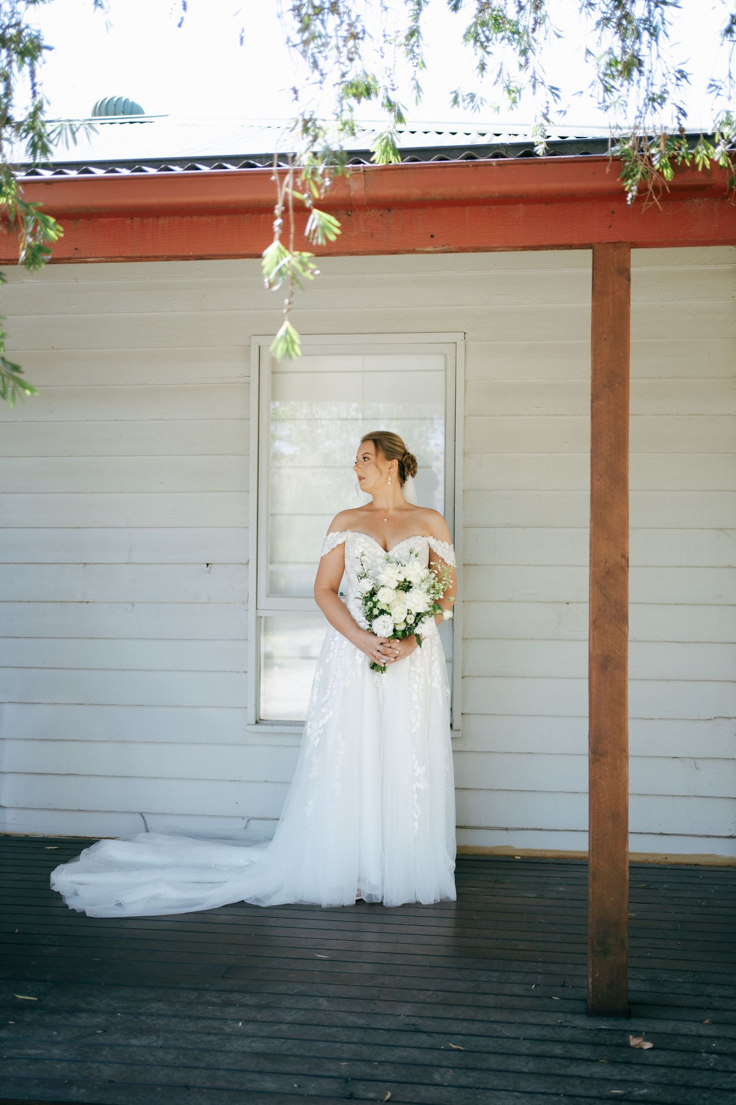 Bride in off-shoulder gown poses with bouquet against white building with wooden porch.