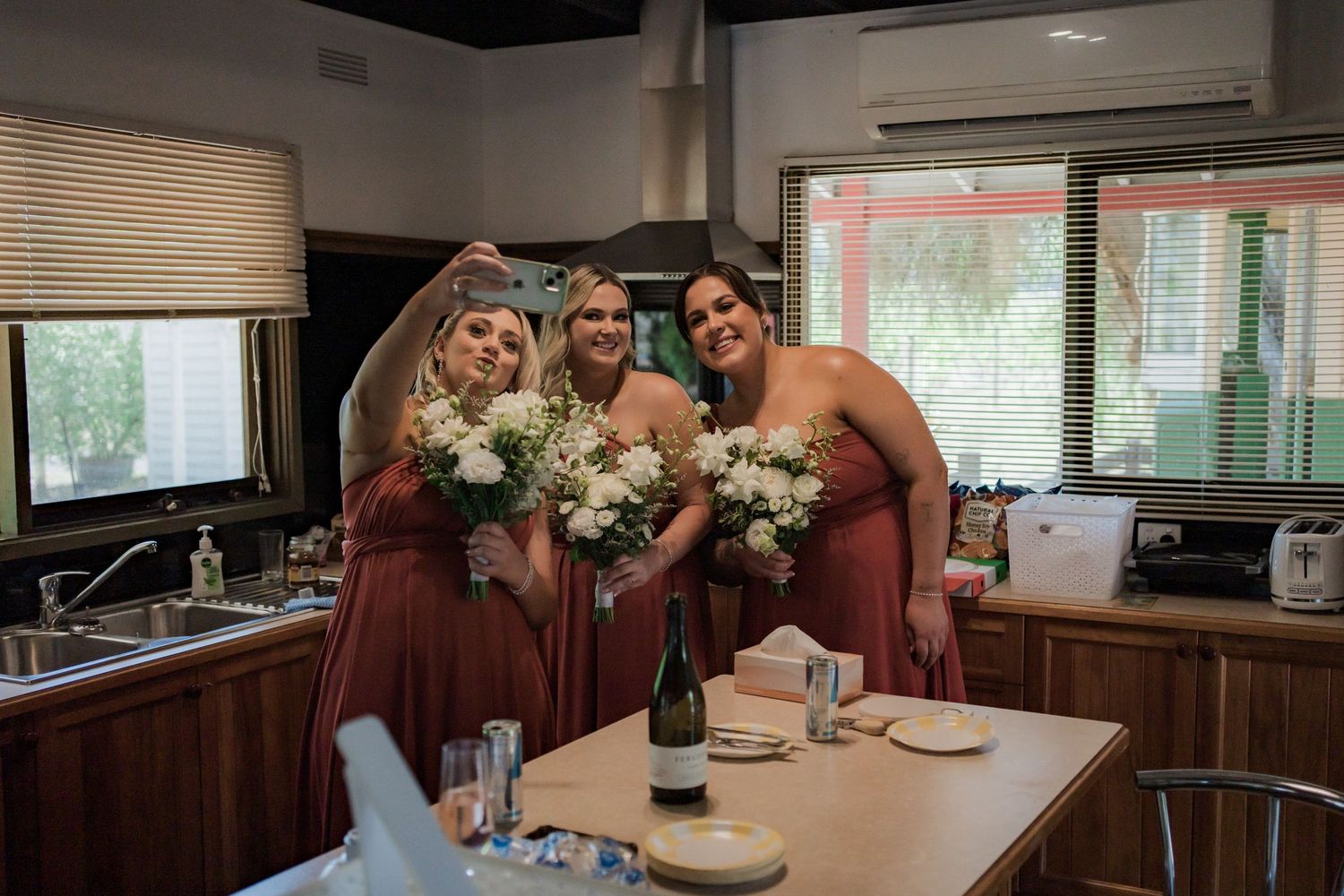 Three bridesmaids in burgundy dresses hold white bouquets while posing in a kitchen setting.