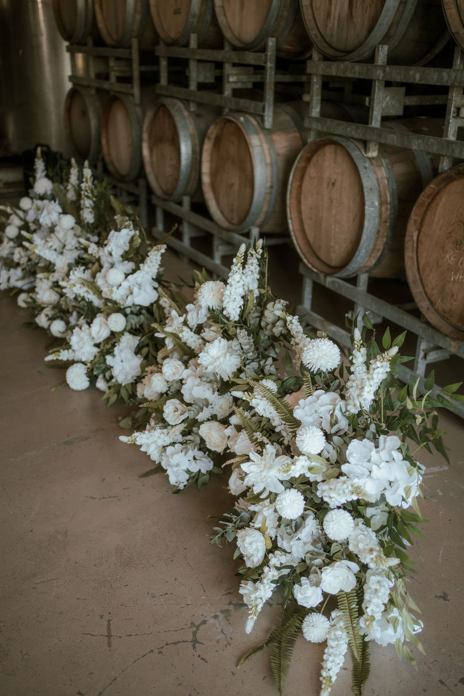 White floral arrangements displayed against wooden wine barrels in a cellar setting.