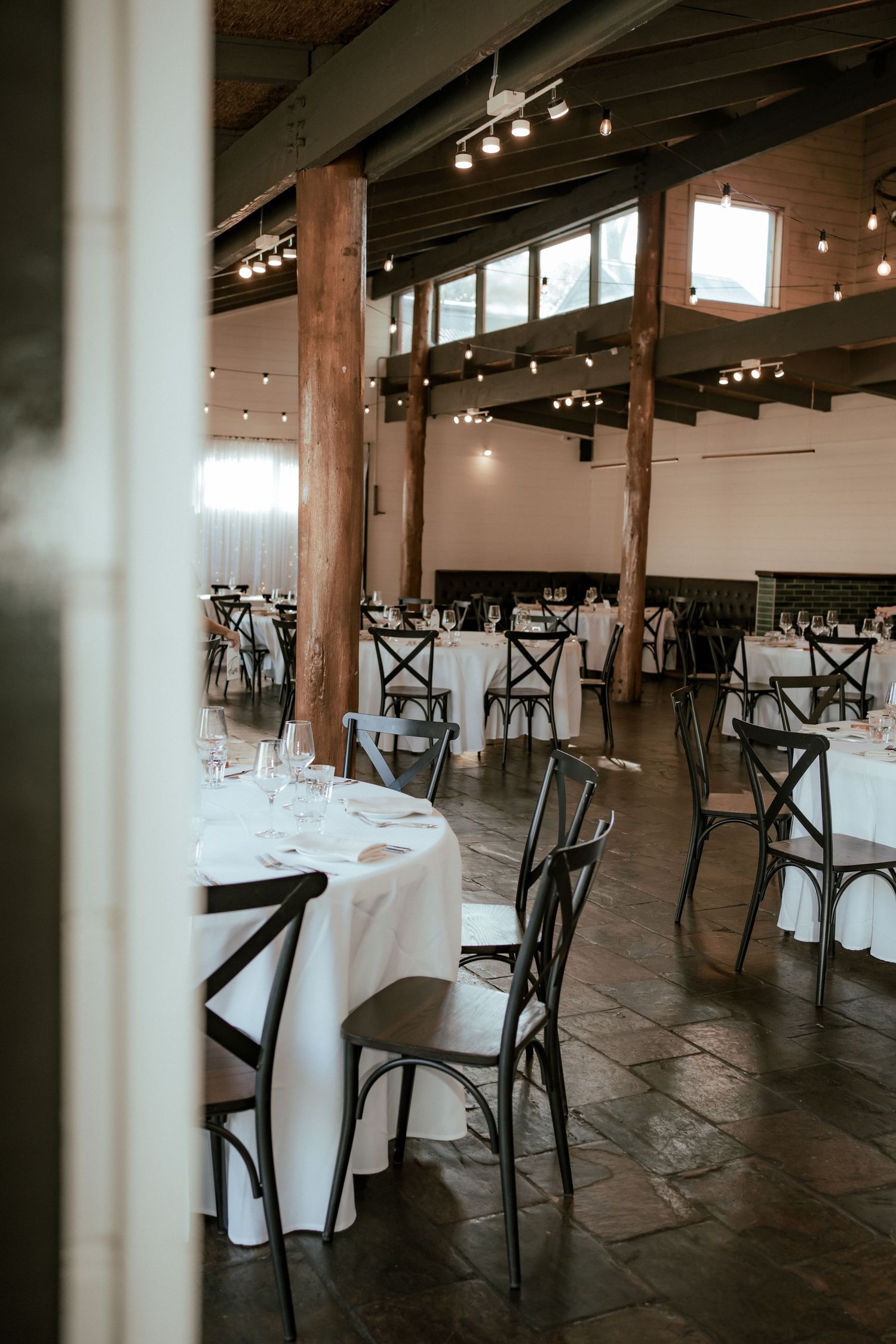 Modern reception venue with white tablecloths and cross-back chairs under industrial lighting.