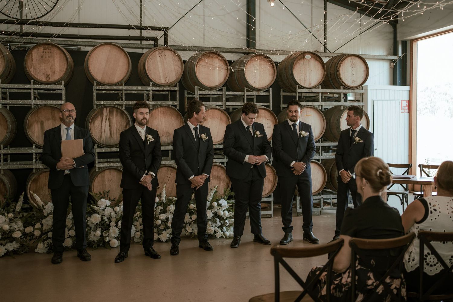 Groomsmen in black suits lined up in front of wine barrels at a winery wedding.