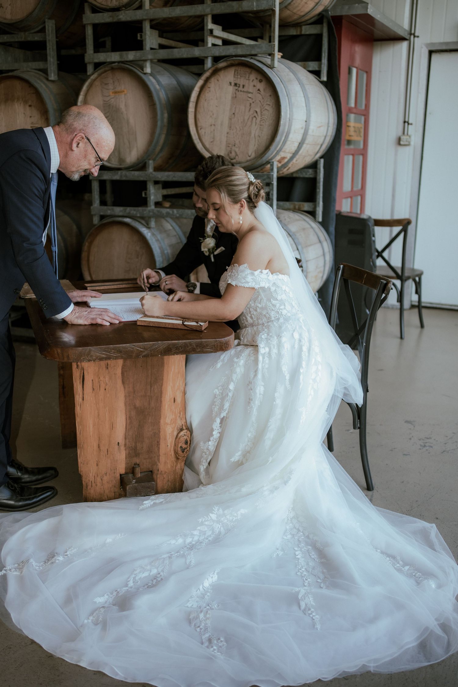 Bride in white lace gown signing documents in front of wine barrels.