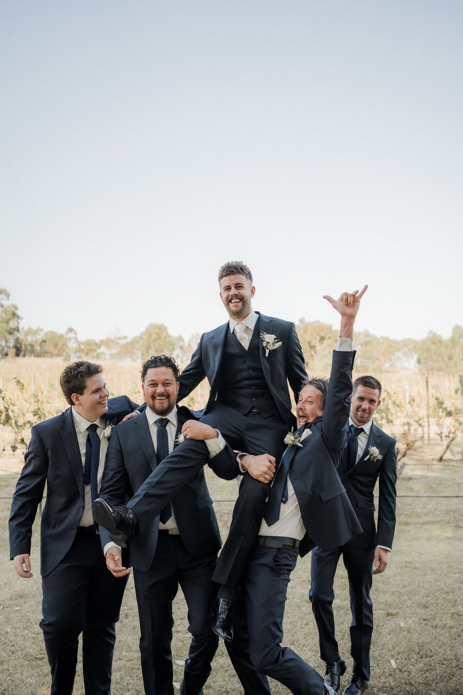 Wedding party lifts groom in playful pose among vineyard rows.