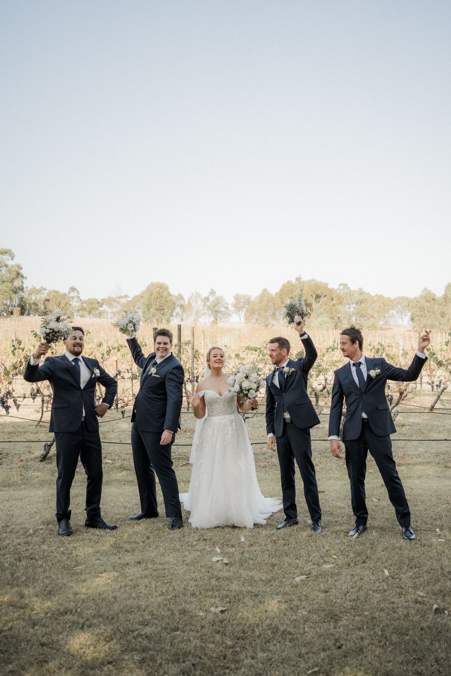 Wedding party celebrates with raised arms in rustic vineyard setting.