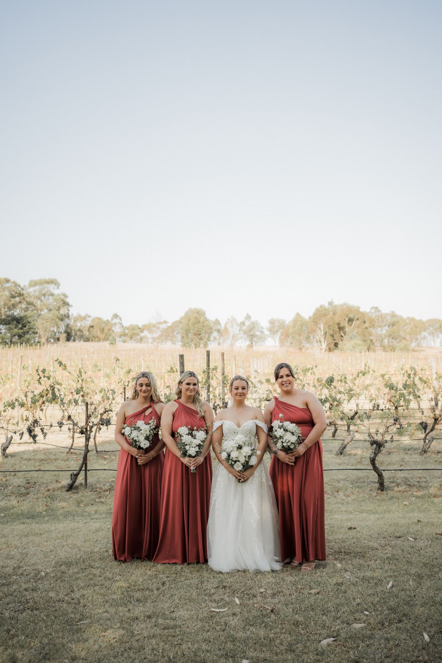 Bridesmaids in burgundy dresses pose with bride among vineyard rows.