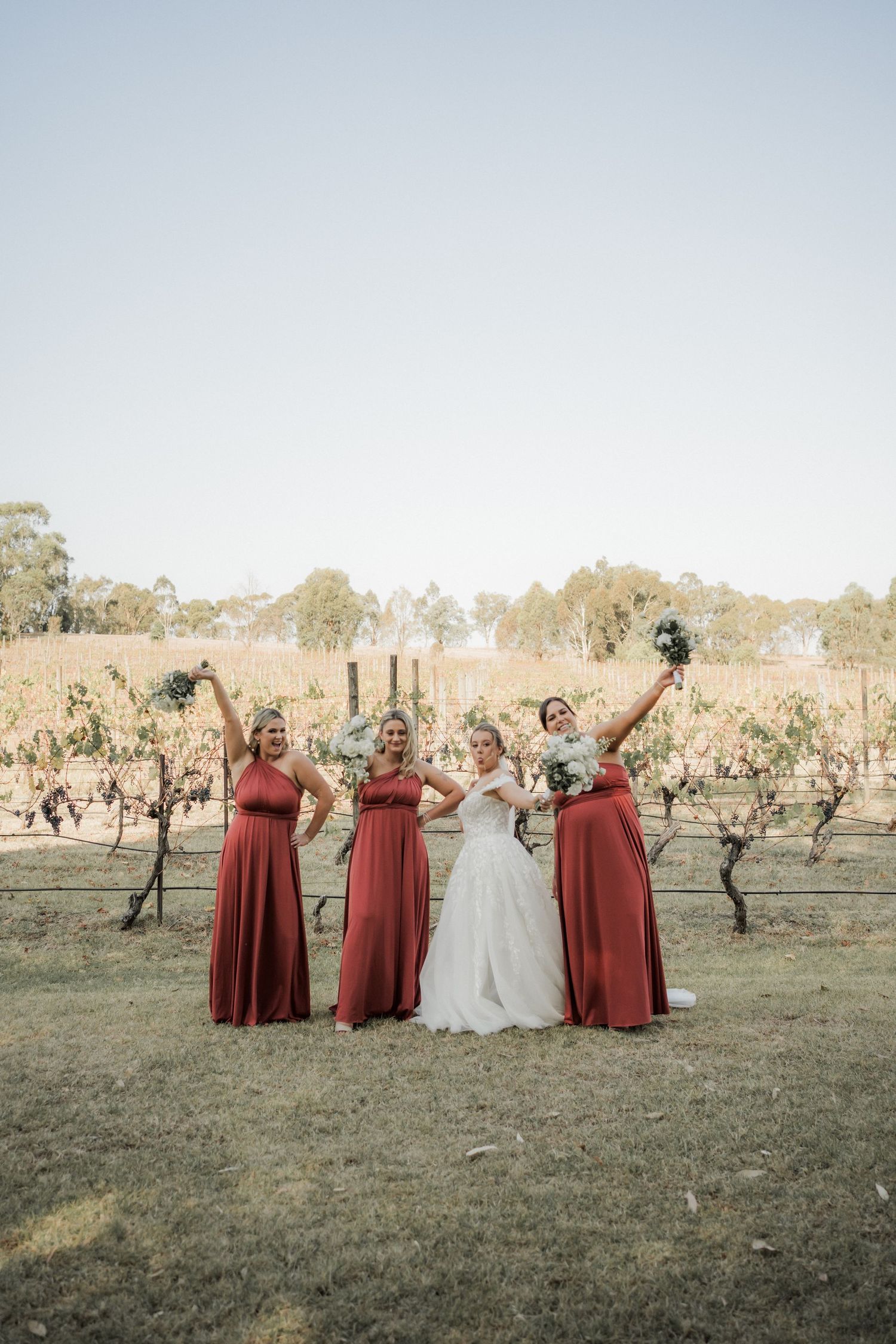 Wedding party in burgundy and white dresses raise their arms in celebration at vineyard.