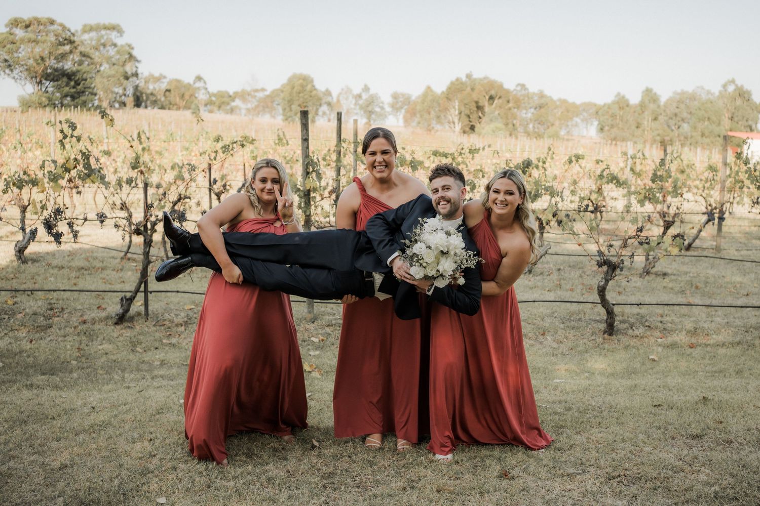 Bridesmaids in burgundy dresses playfully carry bride in vineyard setting.