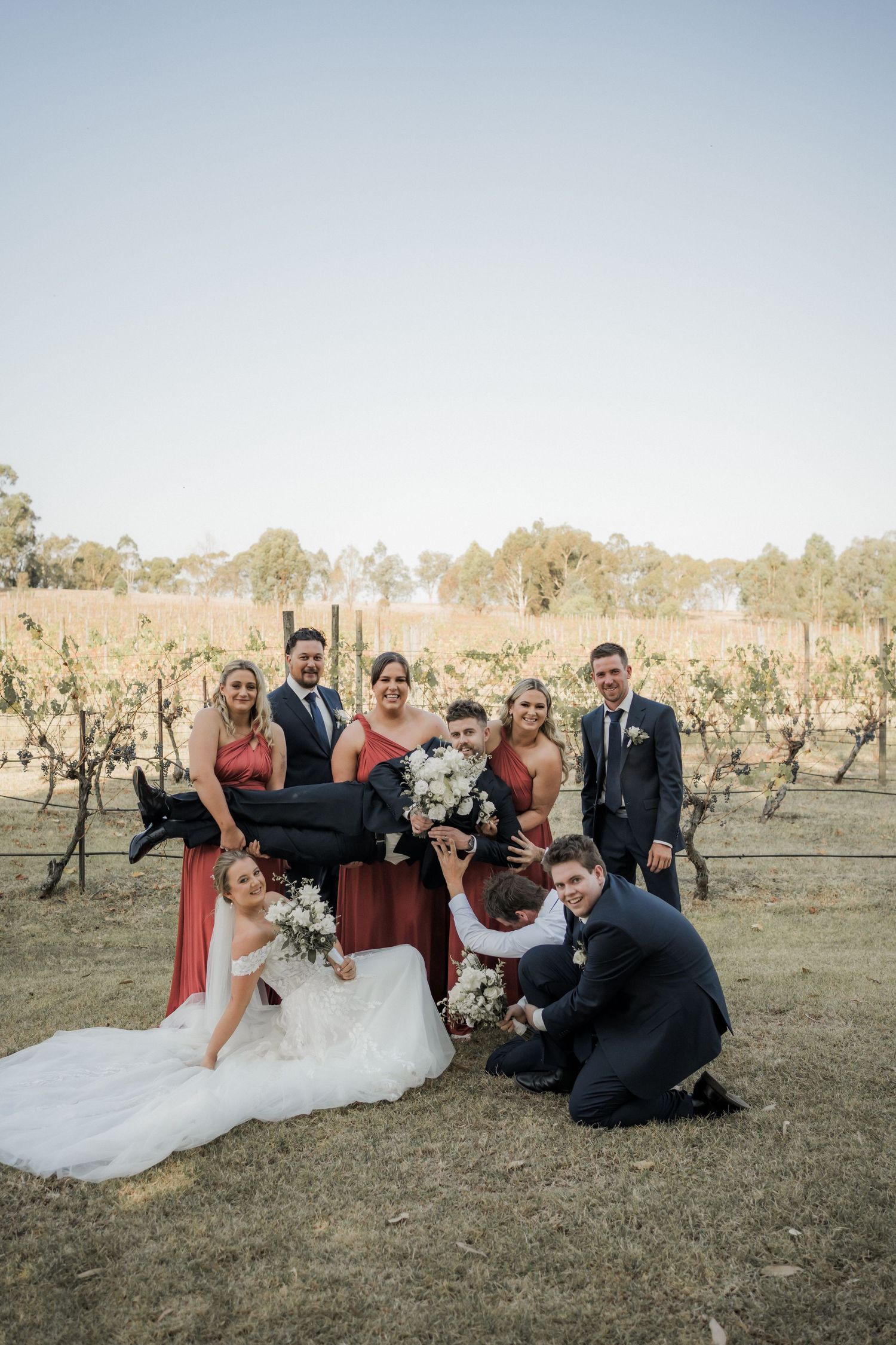 Wedding party poses together in rustic vineyard setting wearing burgundy and black formal attire.