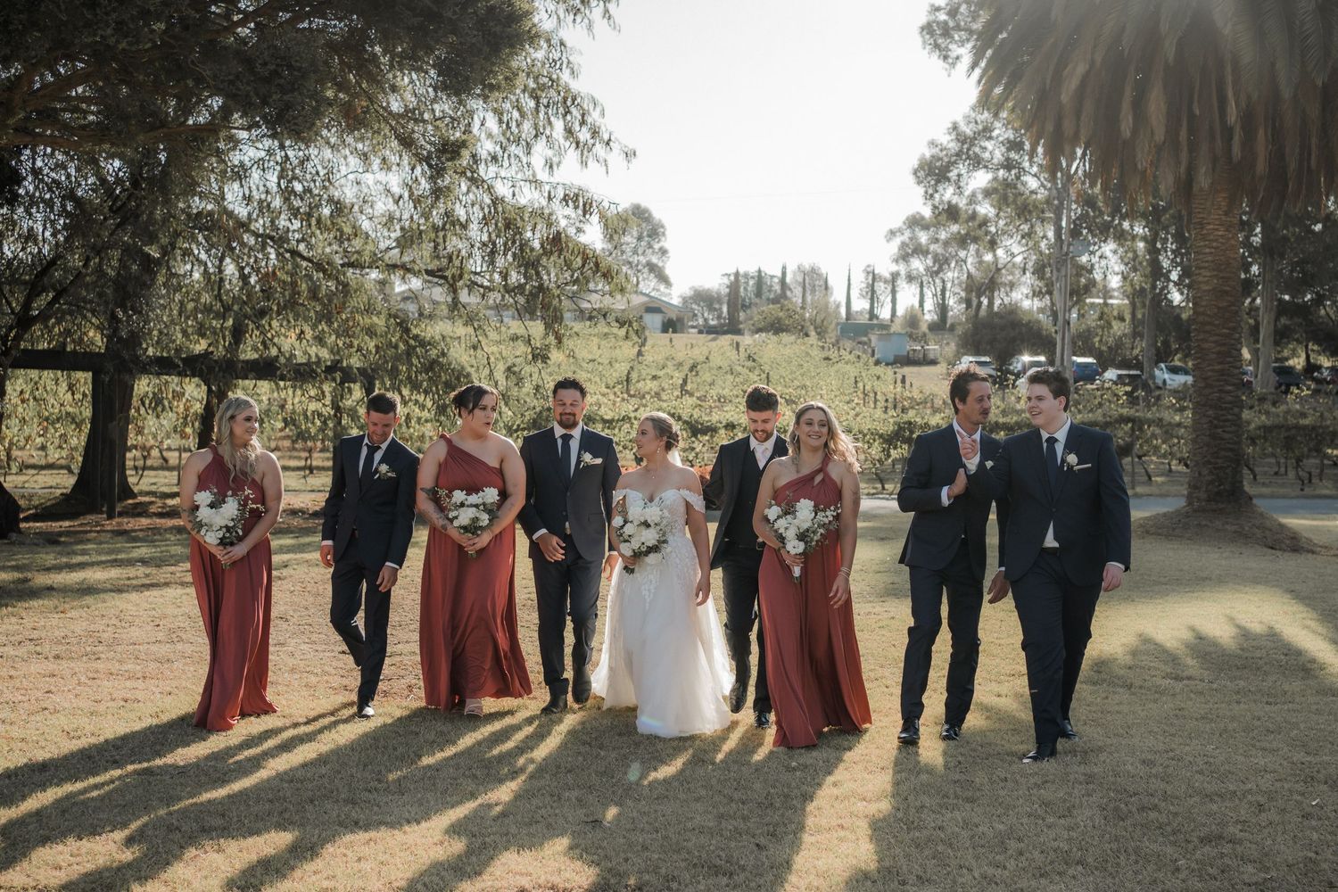 Wedding party walks together on path with vineyard backdrop in burgundy and black formal wear.
