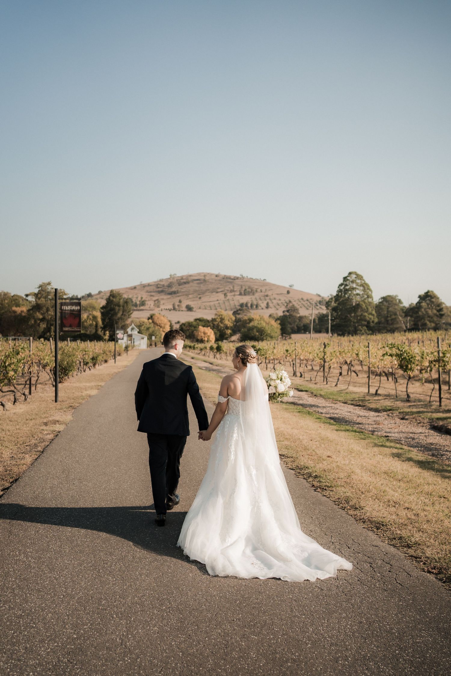 Bride and groom walk down vineyard path with rolling hills in background.