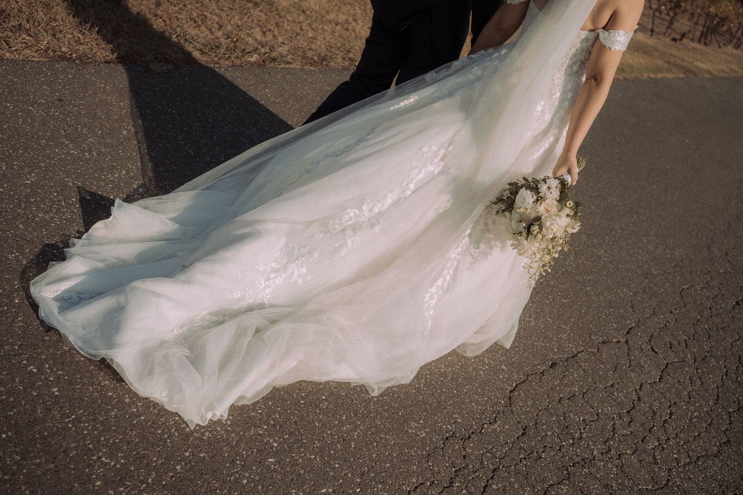 Close-up of flowing white wedding dress train on asphalt path.