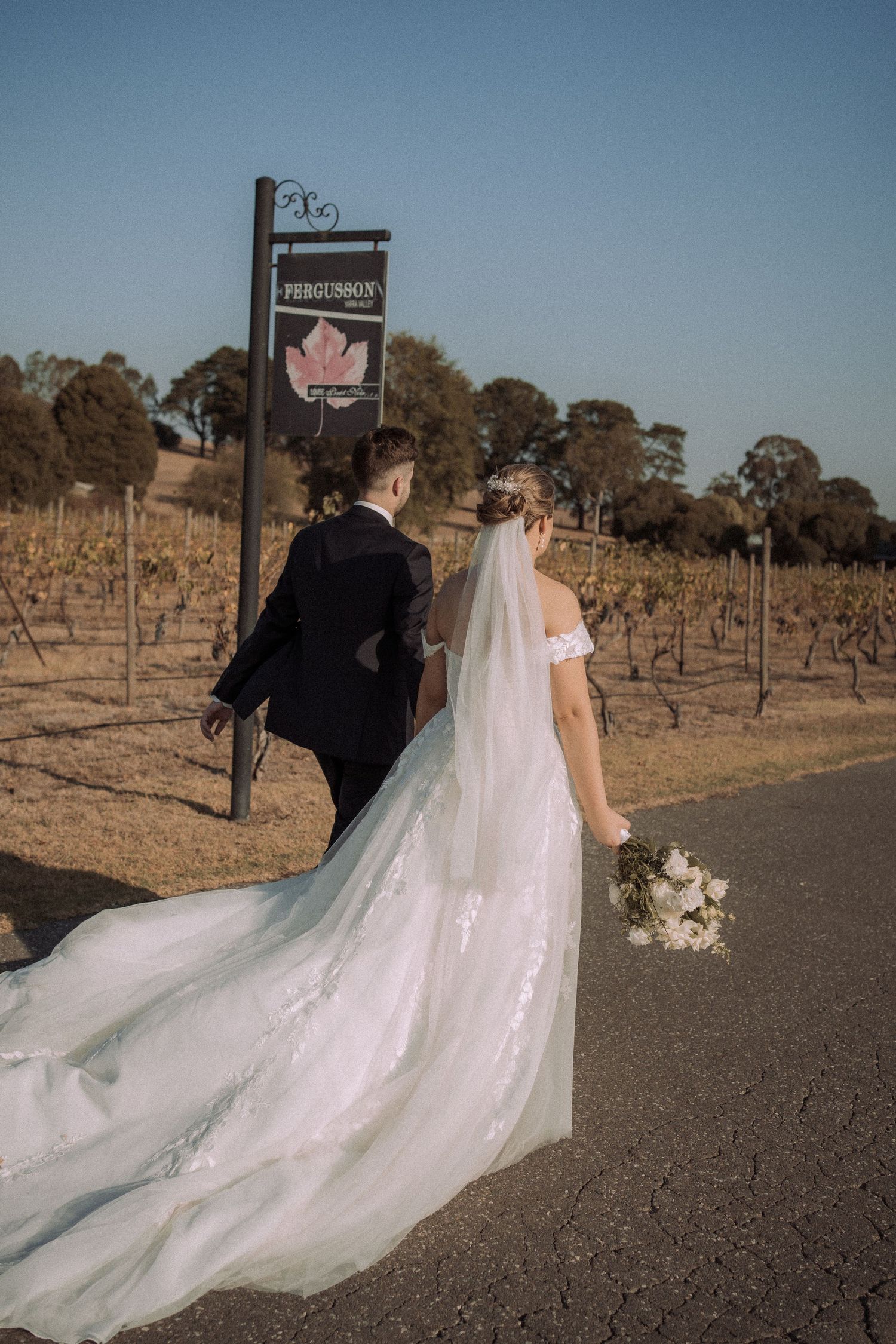 Elegant wedding couple walking past vineyard sign with flowing white dress train.