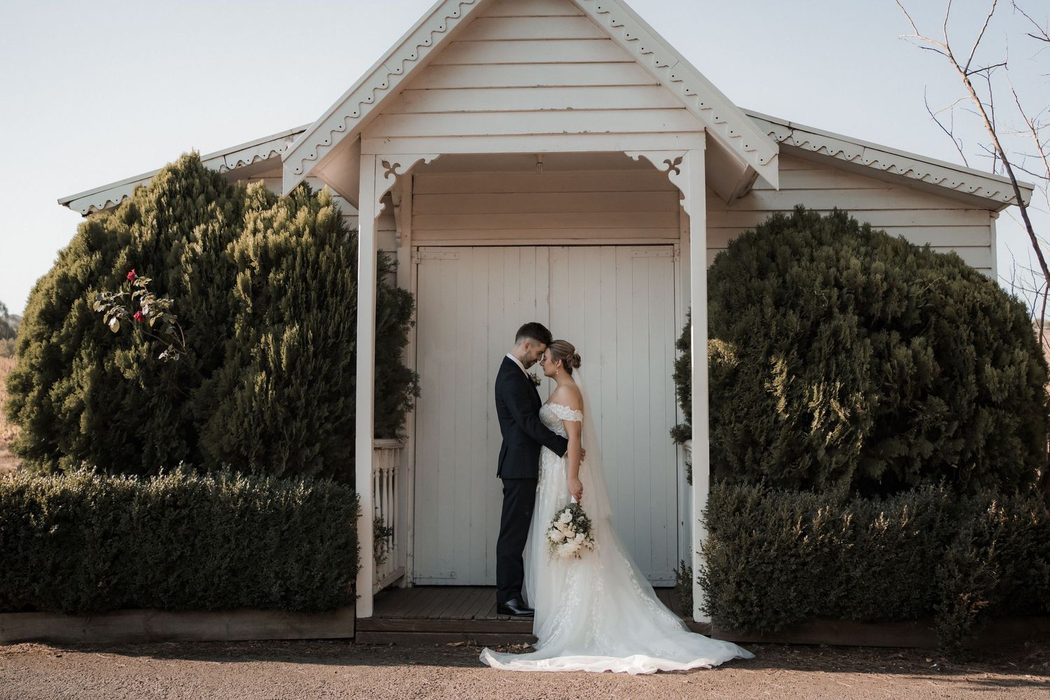 Wedding couple embraces under white gazebo surrounded by lush greenery.