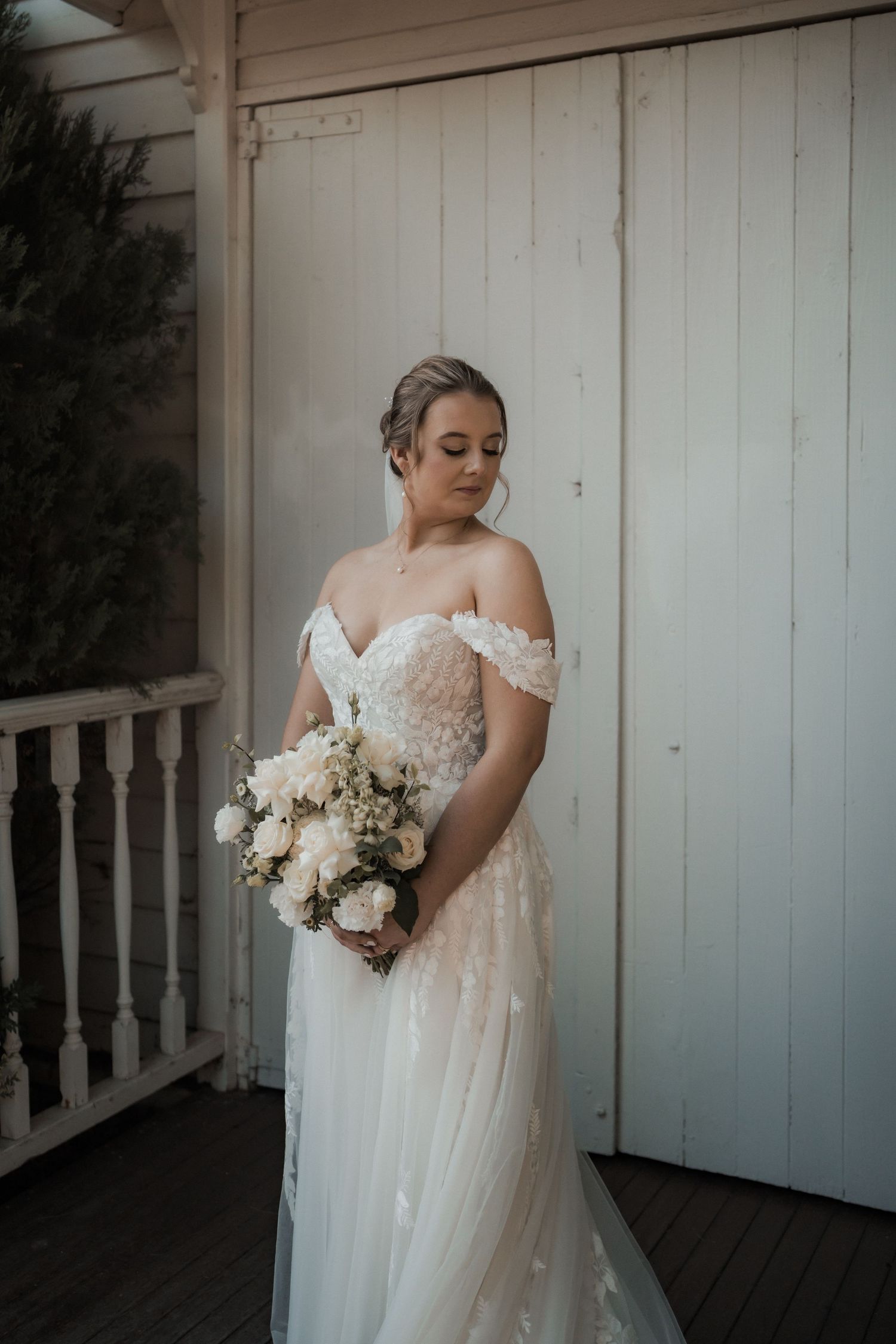 A beautiful off-shoulder wedding gown with delicate bouquet against white wooden backdrop.