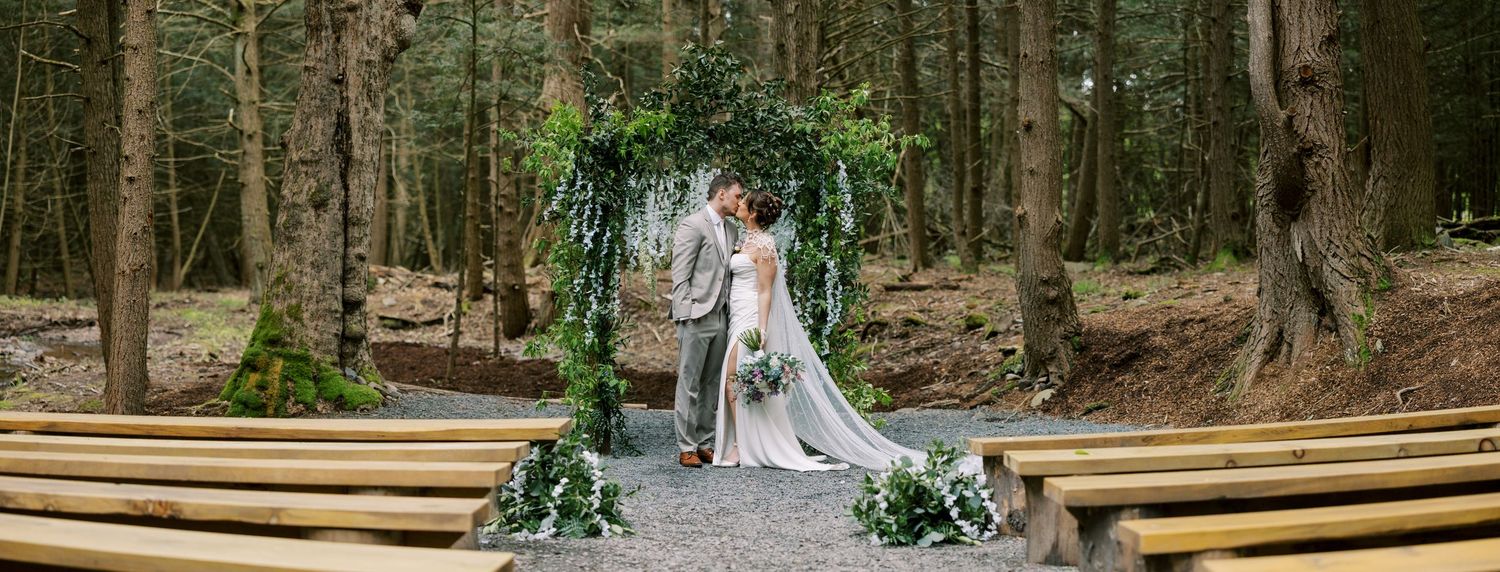 A couple embraces on a path between wooden benches in a forest setting with greenery-decorated wedding arch at Windham Manor.