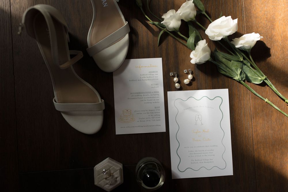 Wedding details flatlay featuring white shoes, invitation cards, and roses on a dark wooden surface.