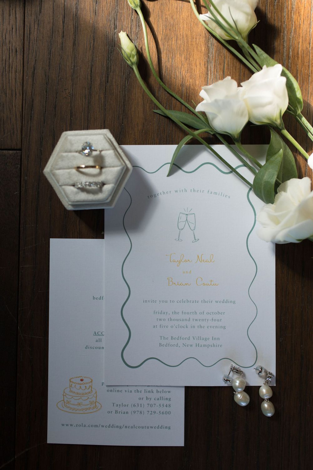 Wedding invitation with white roses, jewelry, and a hexagonal ring box on dark wooden surface.