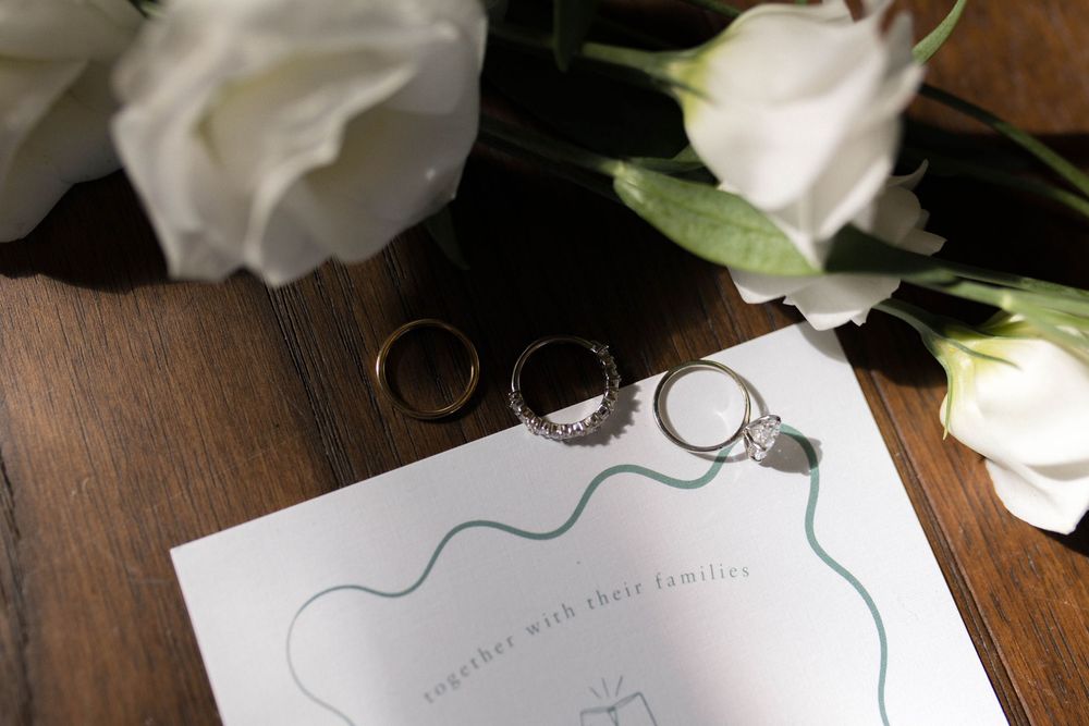 Wedding rings displayed on invitation paper with white roses on wooden surface.