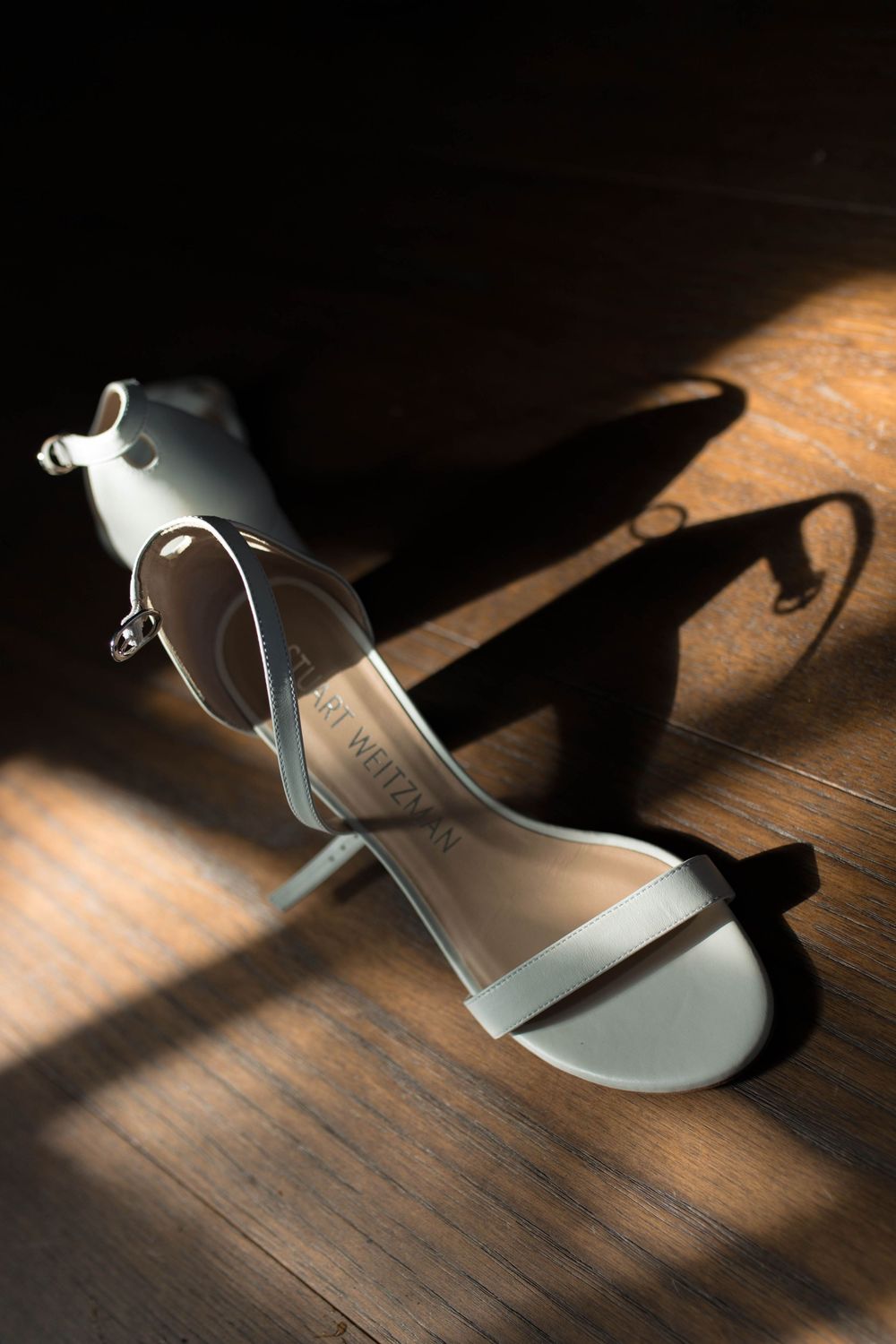 An elegant white bridal sandal casting dramatic shadows on wooden flooring.