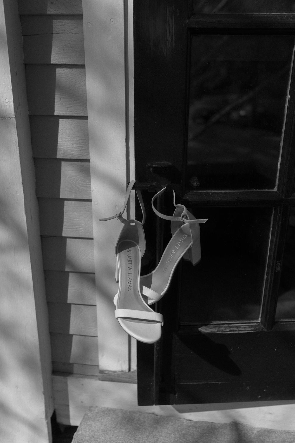 White wedding heels hanging on a dark window frame against white siding in black and white.
