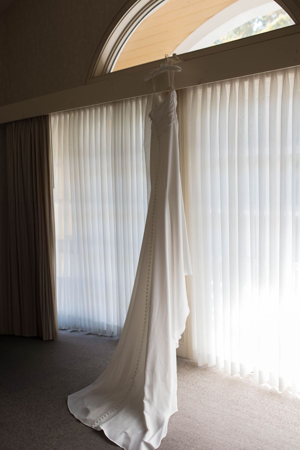 A white wedding dress hangs elegantly against sheer curtains near an arched window.