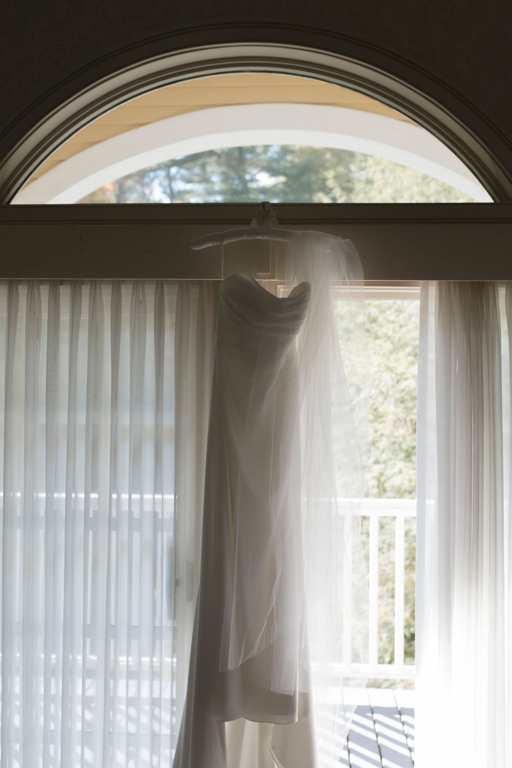 Wedding dress silhouette hanging in arched window with sheer white curtains.