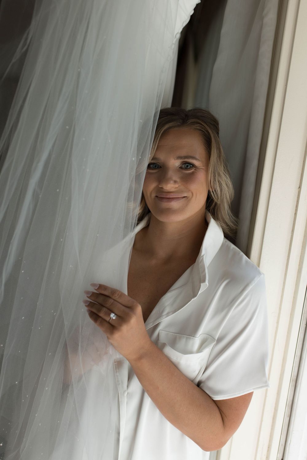 A woman in a white robe smiles while standing near a sheer curtain.