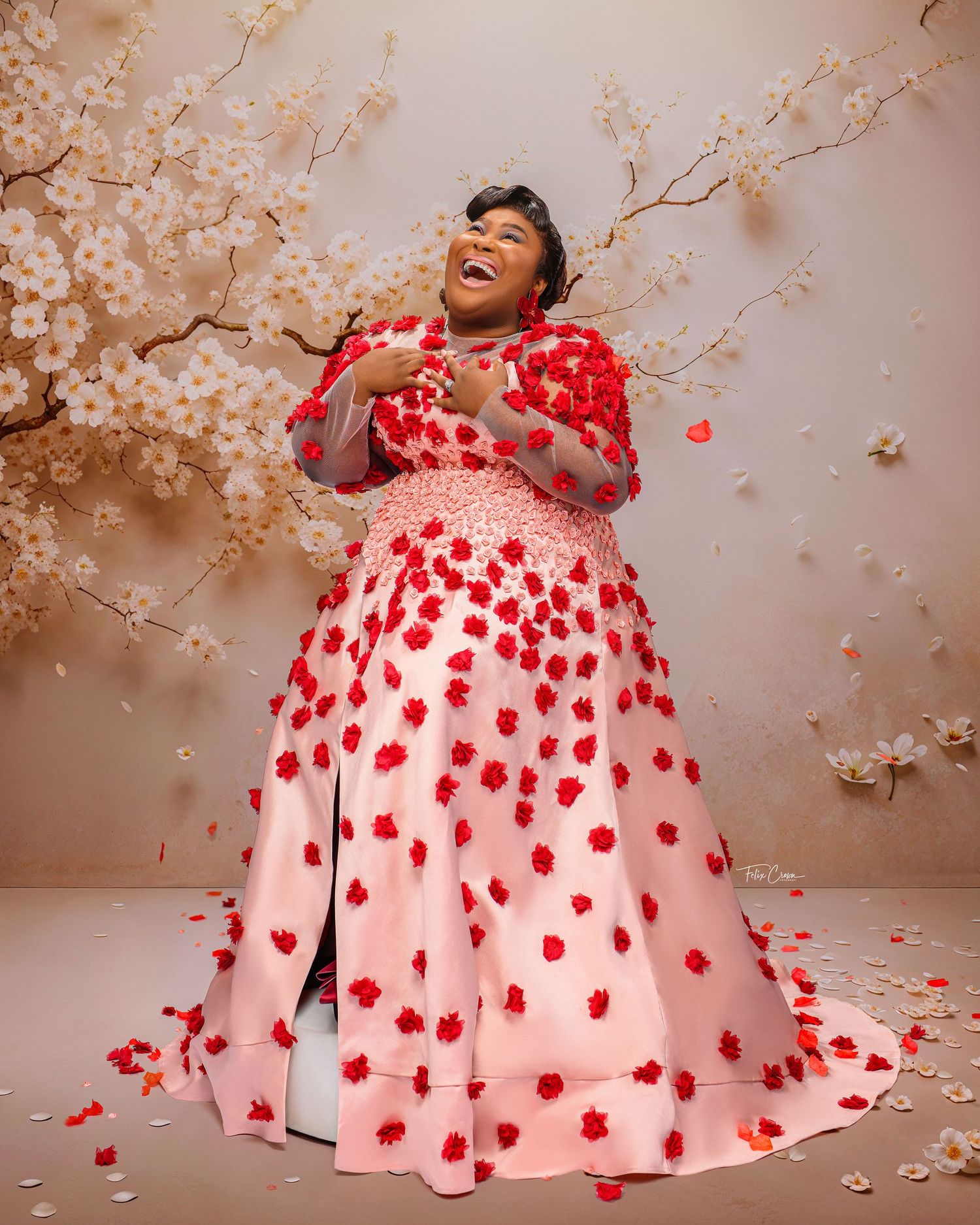 A joyful person in a stunning white and red ball gown poses with cherry blossoms and floating rose petals.