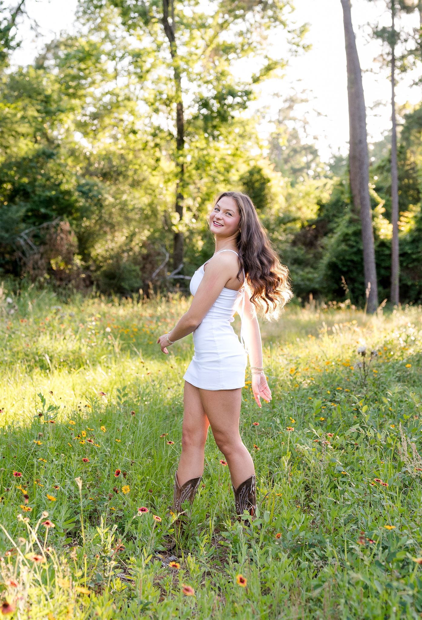 A person wearing a white sundress and brown boots stands in a sunlit meadow surrounded by trees and wildflowers.