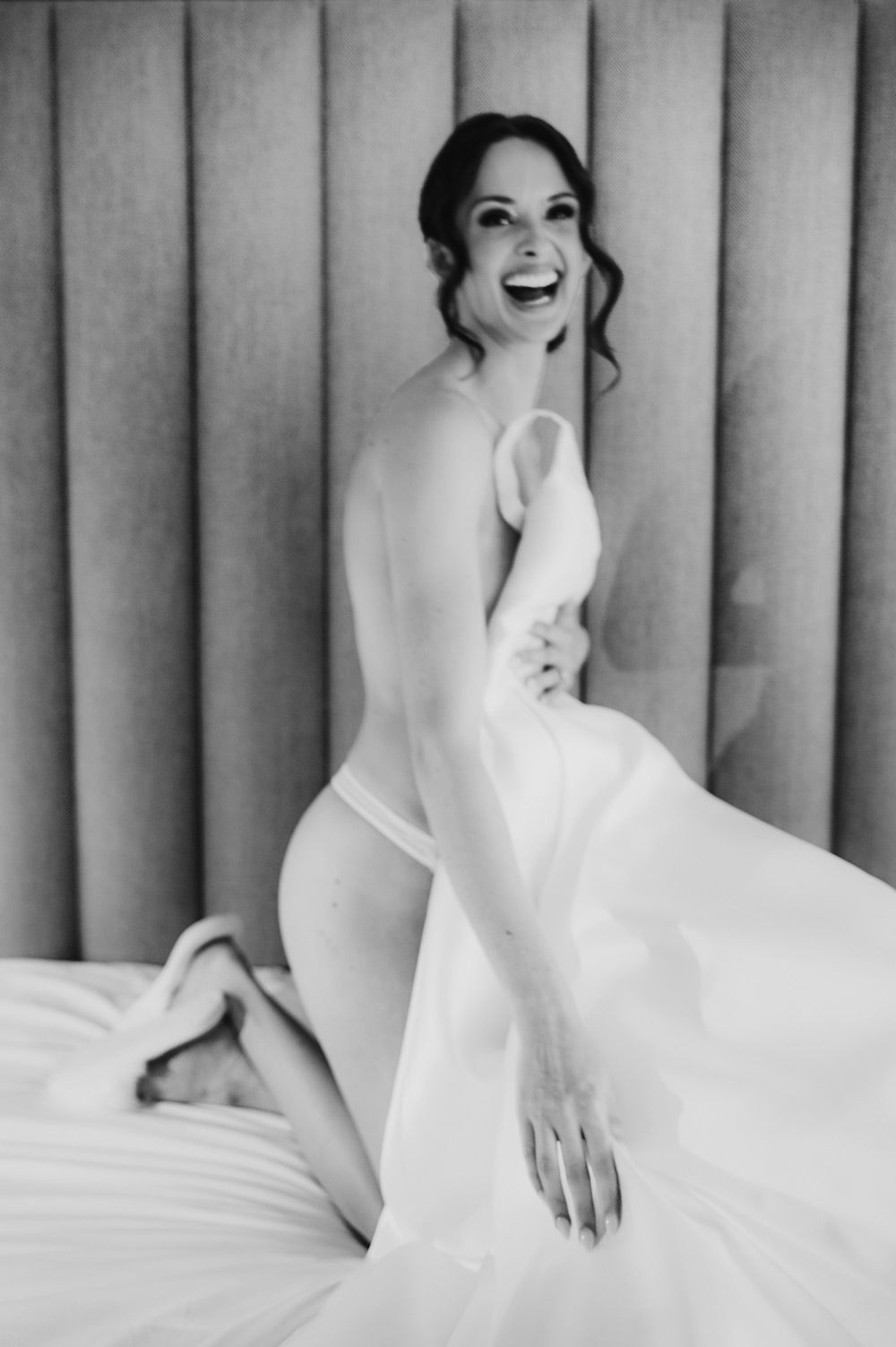 Intimna boudoir scena: žena u svilenoj spavaćici nježno se smije dok gleda preko ramena."