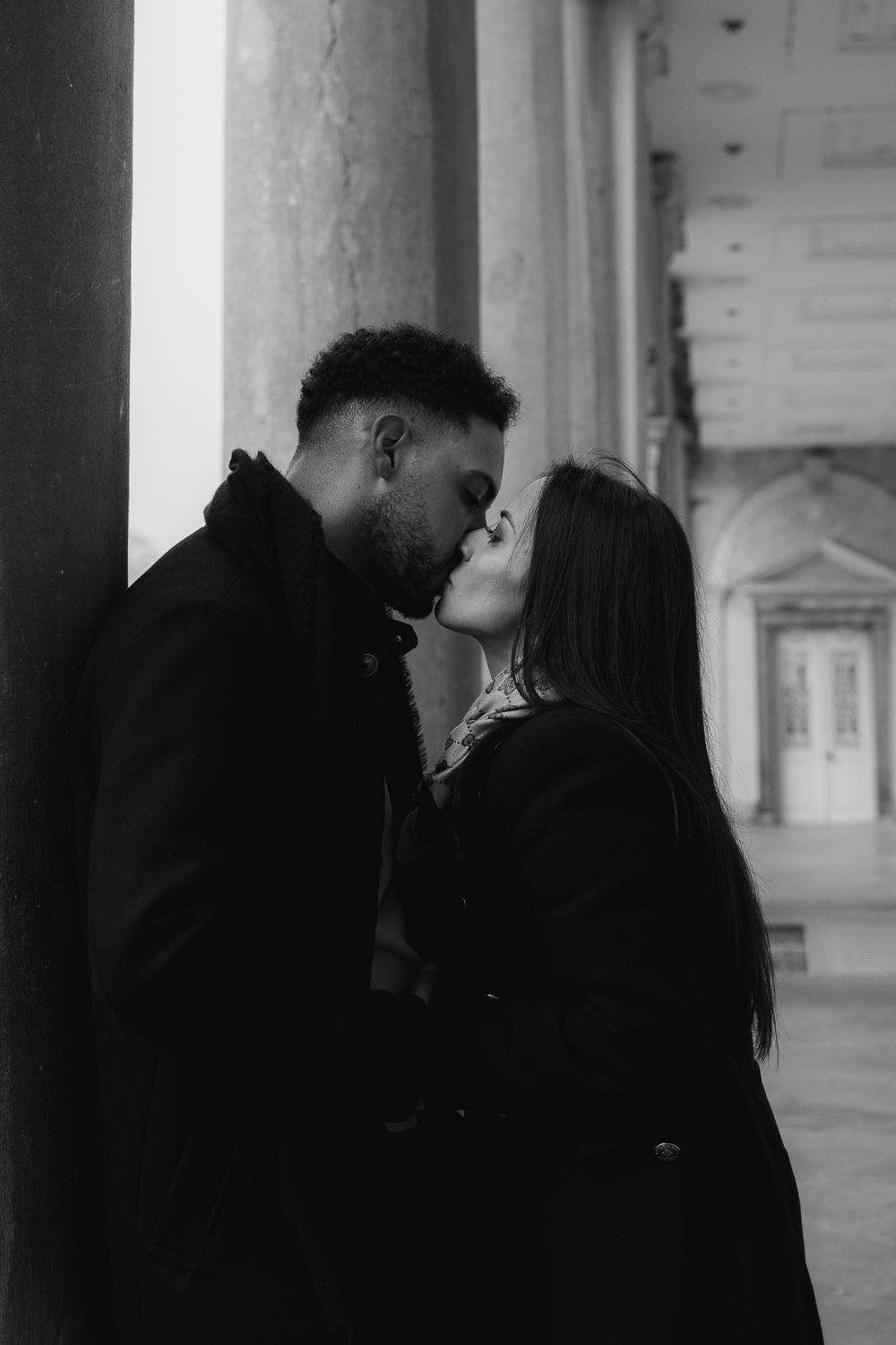 A romantic couple shares an intimate kiss in a black and white photo while standing between columns of a building.